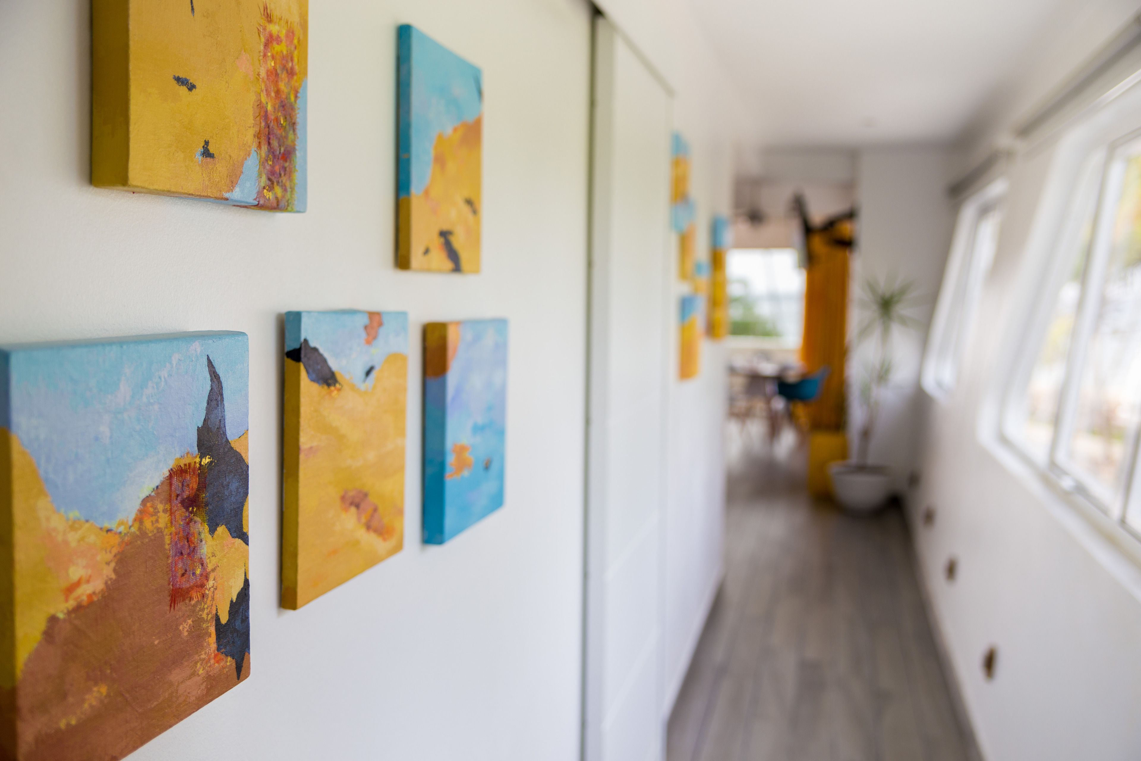 A hallway with abstract paintings on the walls, a wooden floor, and a window on the right.