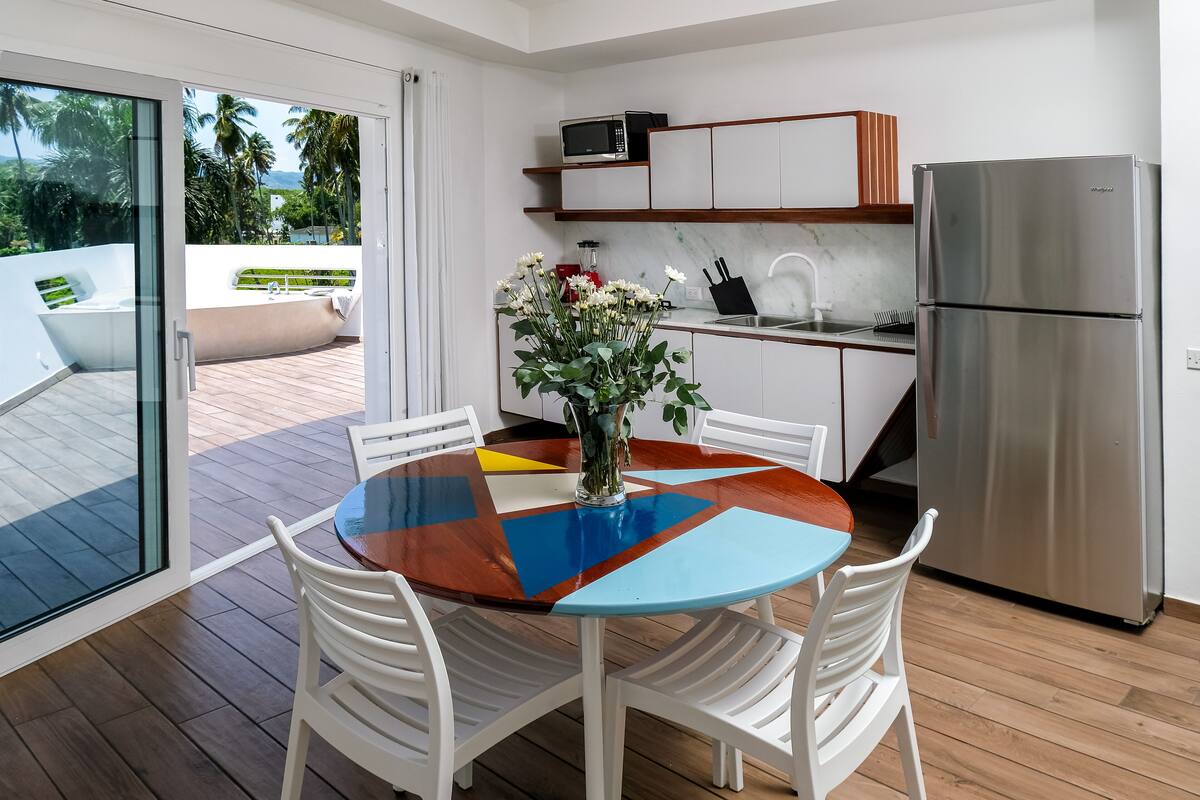 A modern kitchen with a stainless steel refrigerator, a microwave, and a sink. There is a dining table with white chairs and a colorful tablecloth. A vase with flowers is on the table.