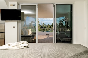 Habitación de hotel con cama, televisor y puerta corrediza de vidrio que da a un balcón con vistas a palmeras y una piscina.