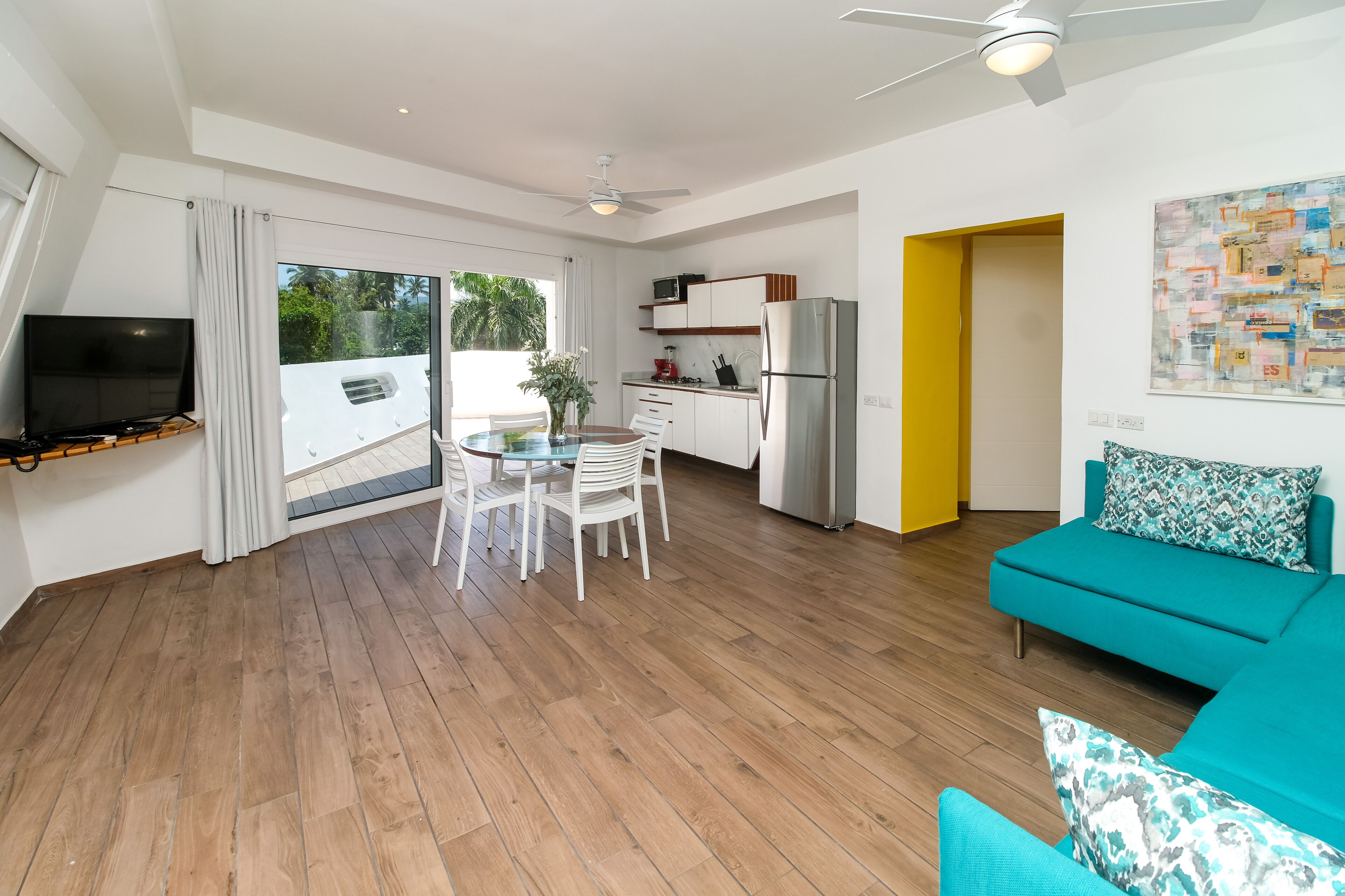 A modern living room with a teal sofa, a dining table with chairs, a kitchen area with a refrigerator, and a TV mounted on the wall.