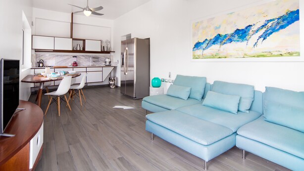 A modern living room with a light blue sectional sofa, a dining area with a wooden table and chairs, a kitchen with white cabinets and stainless steel appliances, and a ceiling fan. - Puerto Plaza Las Terrenas (Las Terrenas)