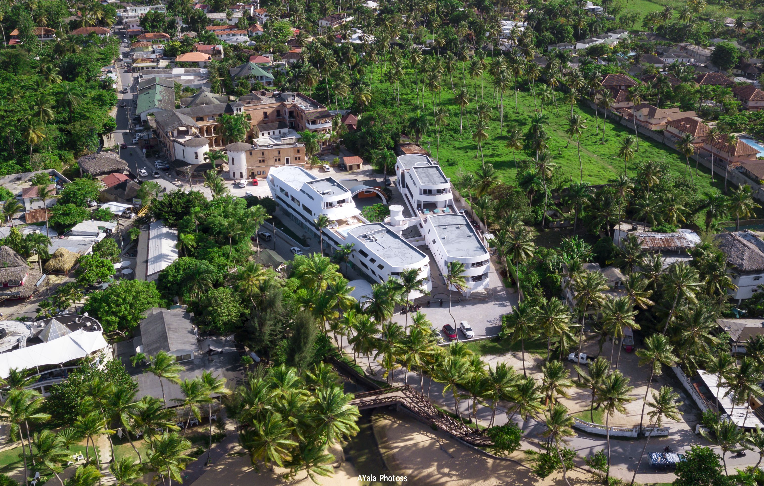 Vista aérea de un pueblo costero con un edificio blanco destacado y techo curvo, rodeado de vegetación exuberante y palmeras.