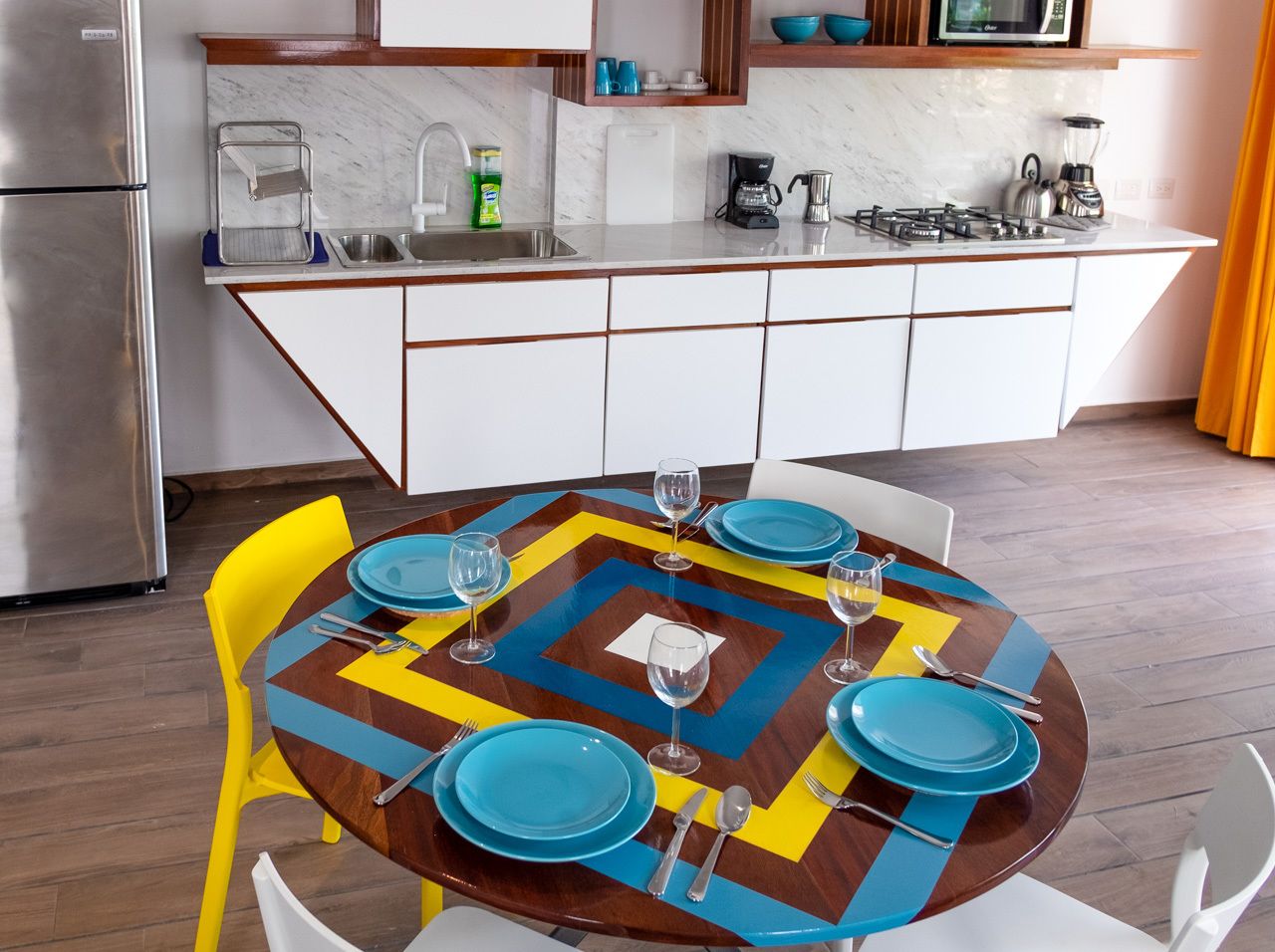 A modern kitchen with a dining table set for six, featuring a colorful geometric tablecloth and matching blue plates.