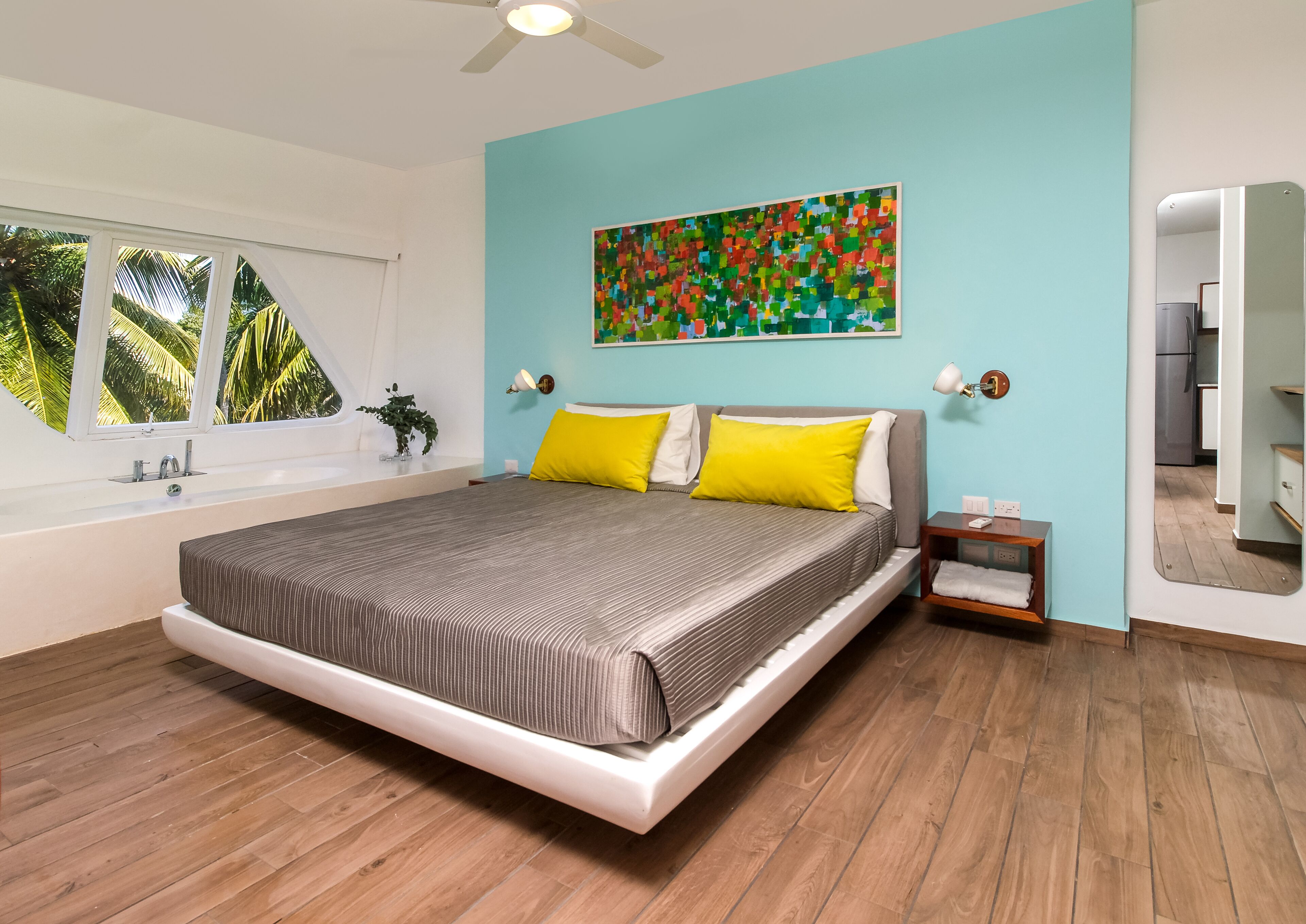 A modern bedroom with a large bed, a window with a view of palm trees, a colorful abstract painting on the wall, and a ceiling fan.