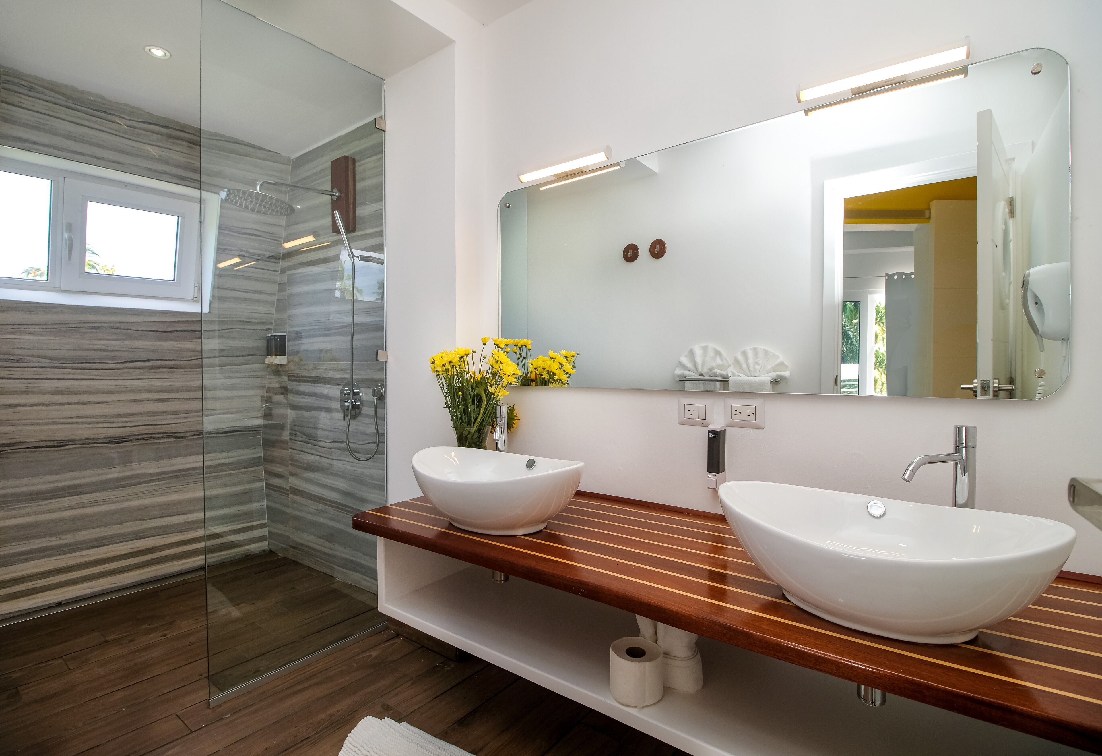 Un baño moderno con cabina de ducha de vidrio, dos lavamanos, un mueble de tocador de madera y un espejo grande.