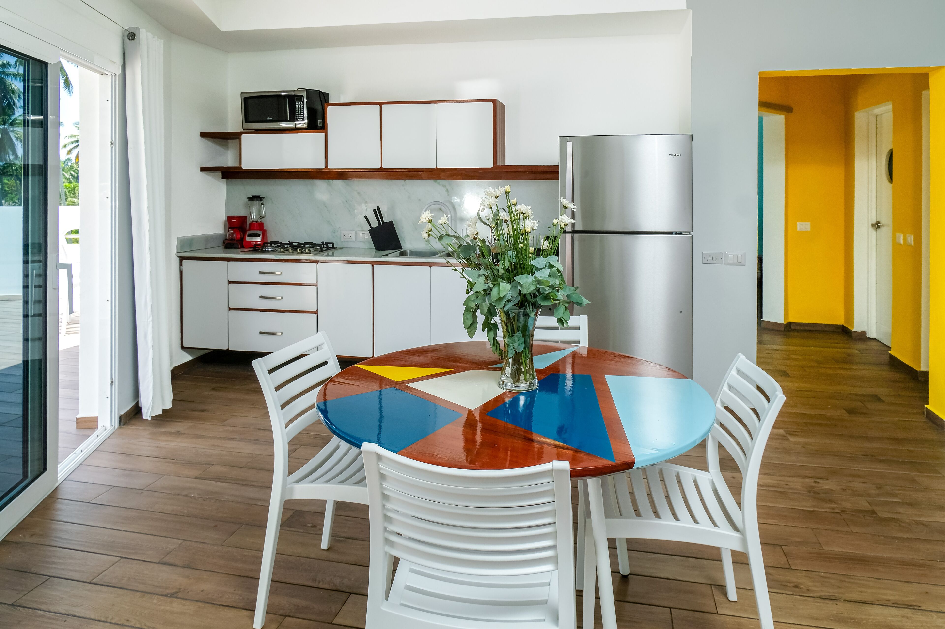 A modern kitchen with white cabinets, a stainless steel refrigerator, and a dining area with a colorful table and white chairs.