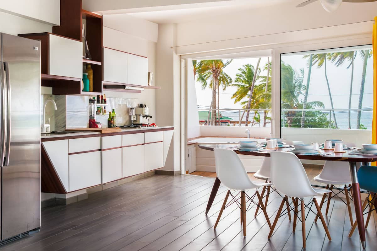 A modern kitchen with white cabinets, a stainless steel refrigerator, and a dining area with white chairs and a table set for four.