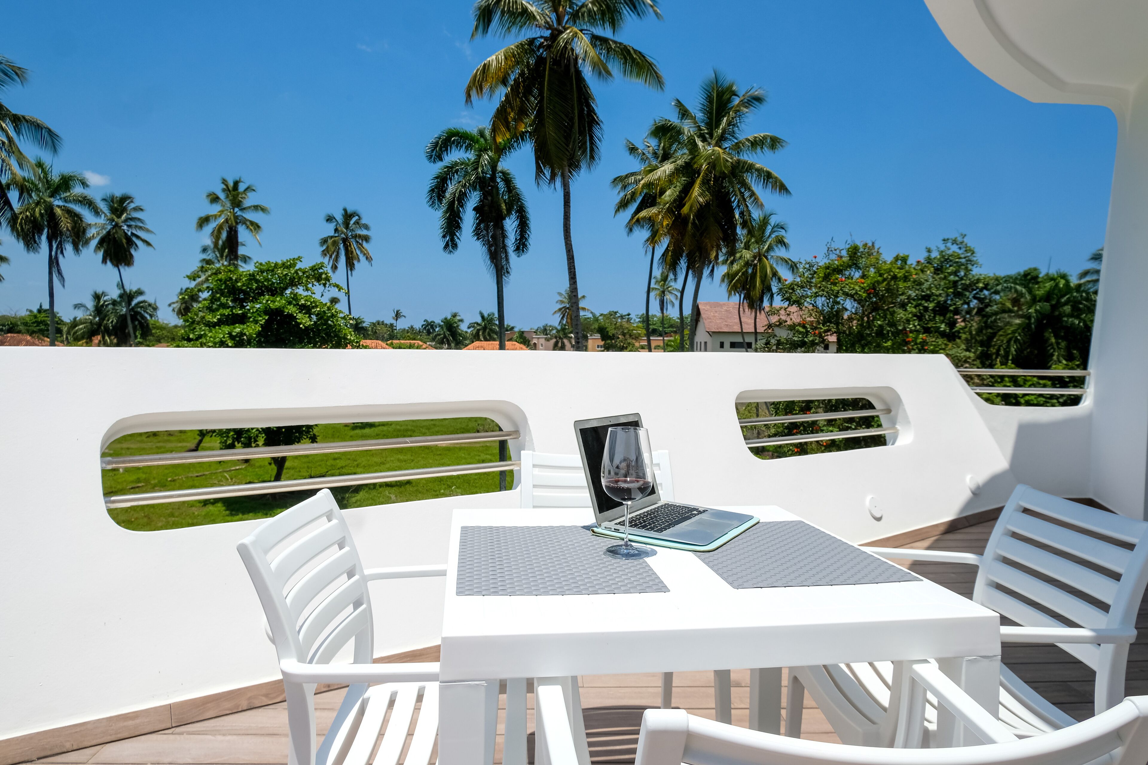 Zona salotto esterna con tavolo, laptop e bicchiere di vino, con vista su un giardino con palme e un cielo azzurro limpido.