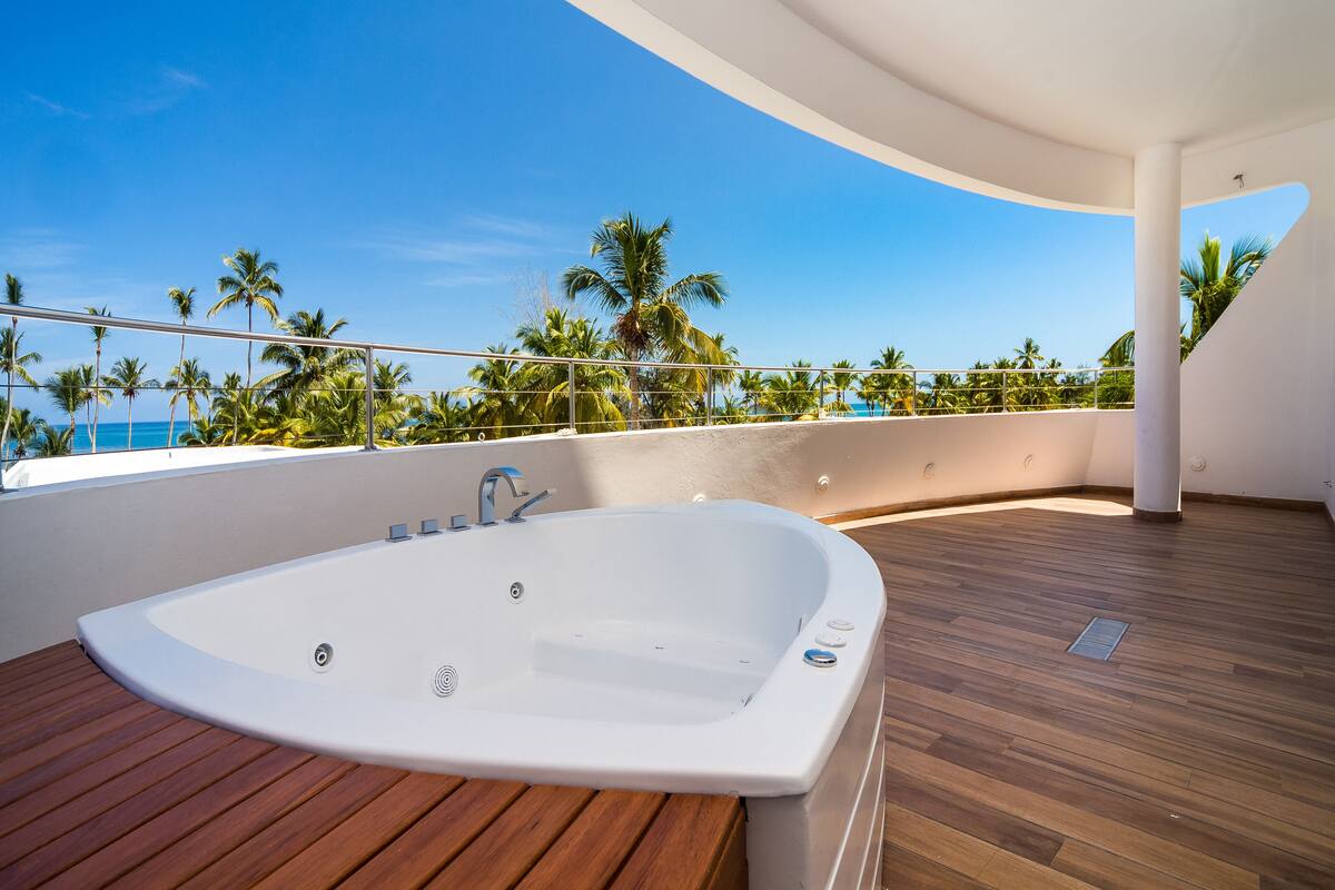 A modern indoor pool area with a large jacuzzi, wooden flooring, and a view of palm trees and the ocean.