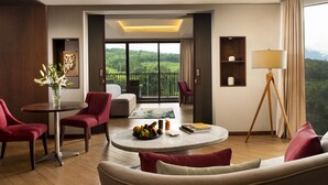 A modern hotel room with a balcony, a round coffee table with fruits, and a view of the greenery outside.
