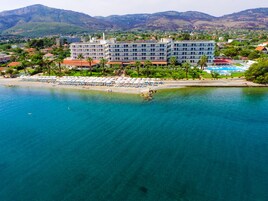 A coastal hotel with multiple floors, a beach with umbrellas, and a mountainous backdrop.