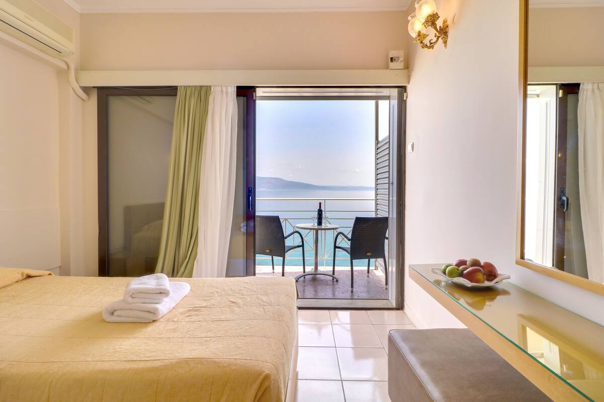 A hotel room with a bed, a glass table with fruit, a balcony with a view of the sea, and a large mirror.
