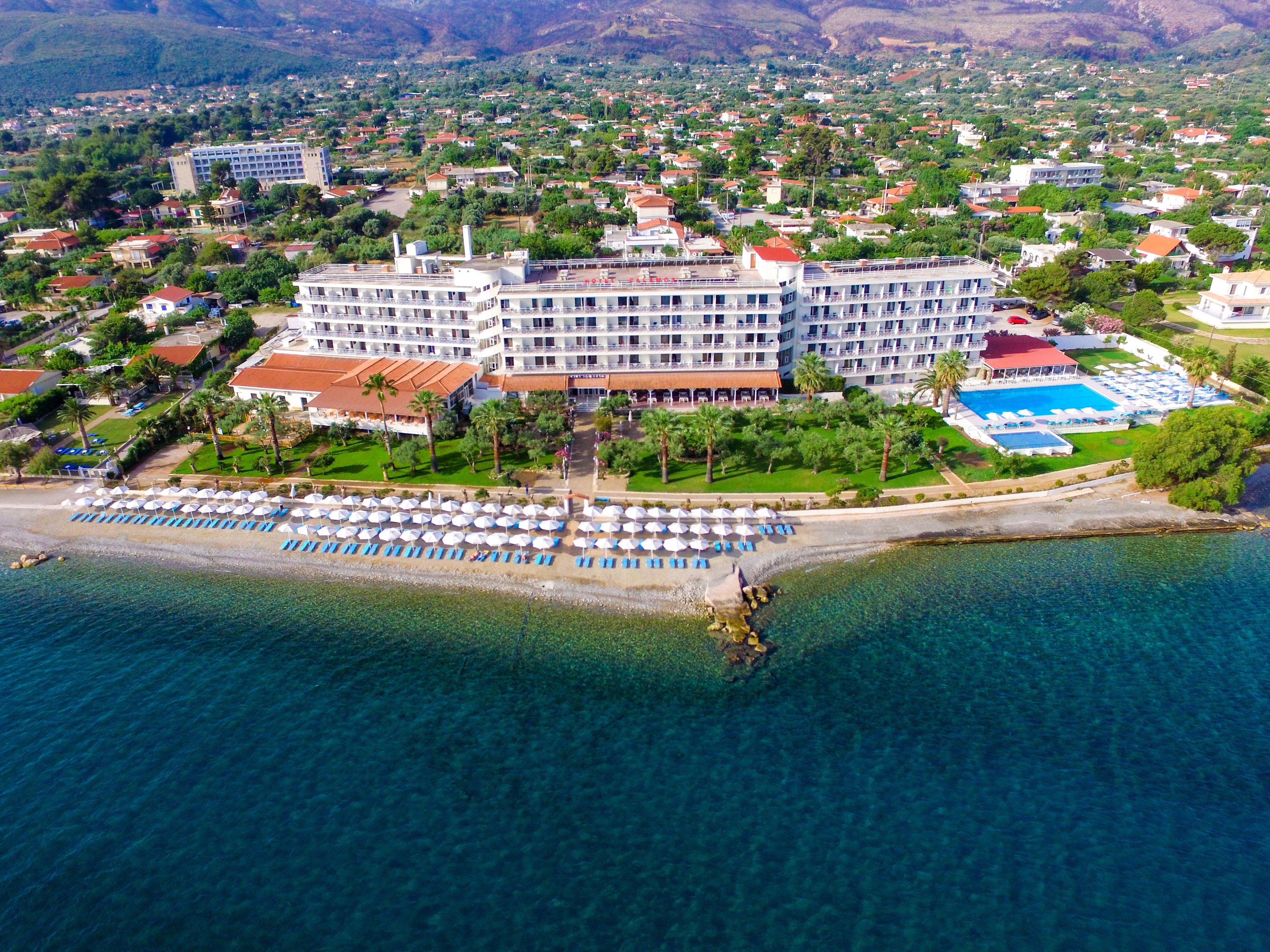 A large hotel with multiple floors, a swimming pool, and a beach area with umbrellas and sun loungers.