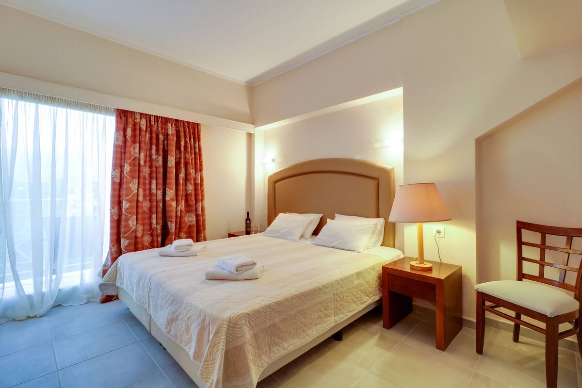A hotel room with a large bed, a nightstand with a lamp, a wooden chair, and a window with curtains.