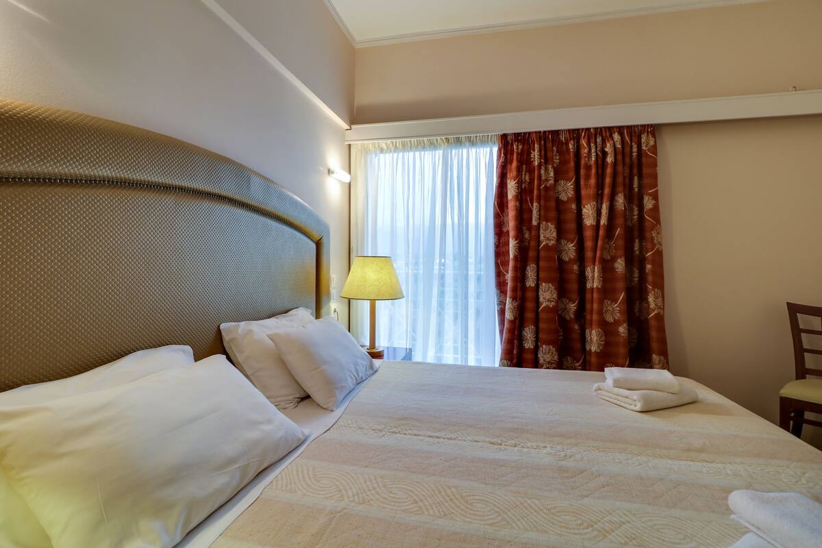 A hotel room with a large bed, a headboard, a lamp, a window with curtains, and a chair.