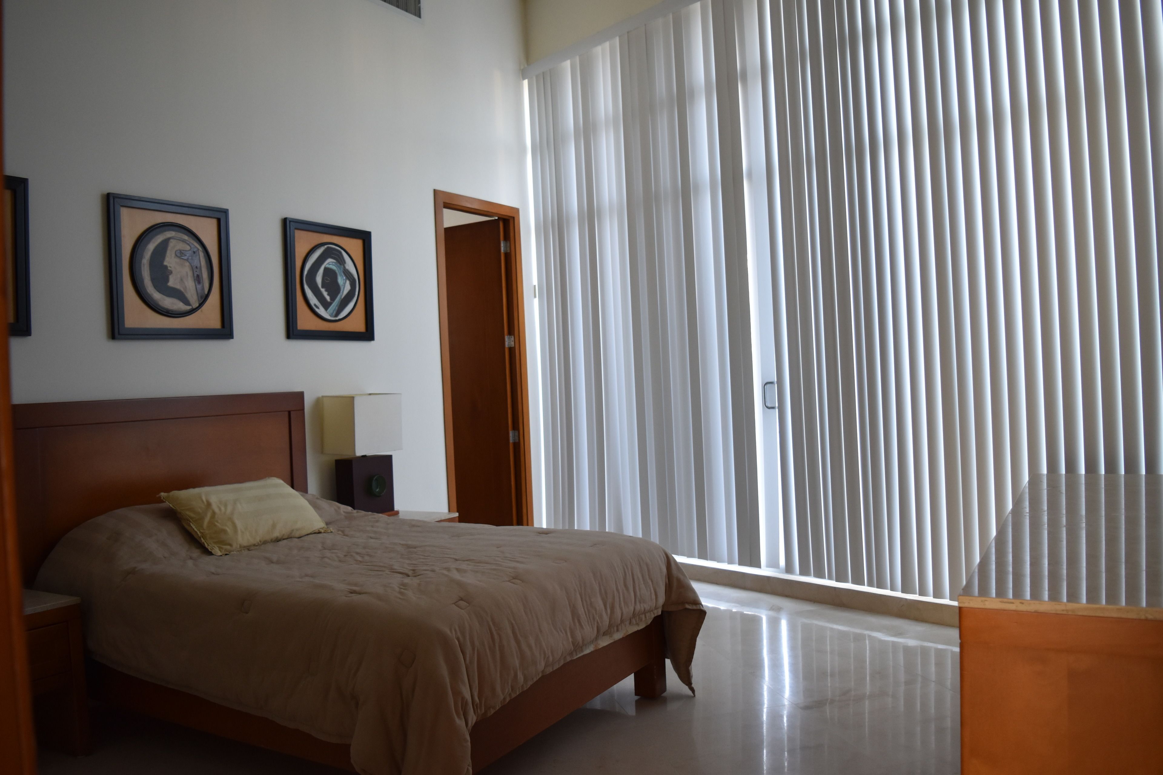 A bedroom with a large bed, a wooden headboard, a nightstand, and a bedside table. There are two framed pictures on the wall and a window with vertical blinds.