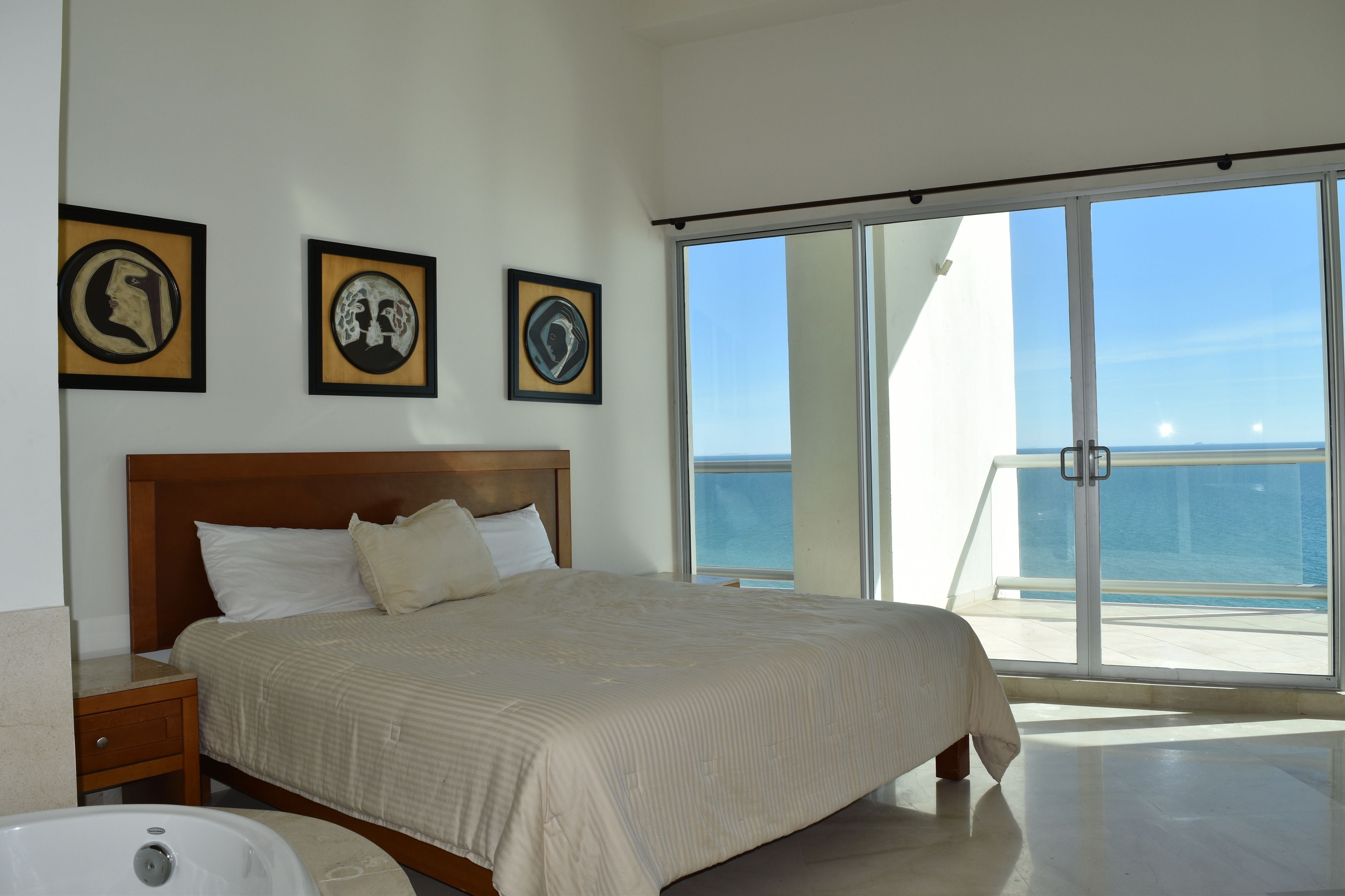 A bedroom with a large bed, a nightstand, a bathtub, and a balcony with a view of the sea.