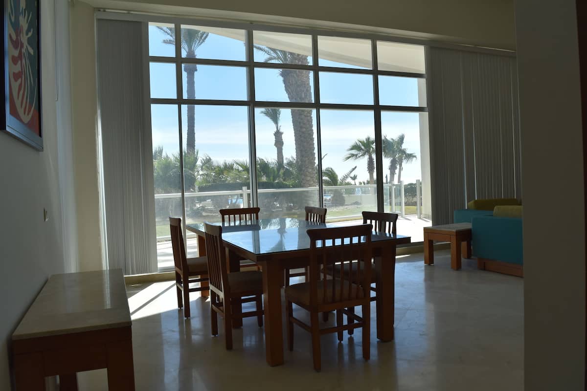 A dining area with a large table and chairs, large windows with a view of palm trees and a clear sky.
