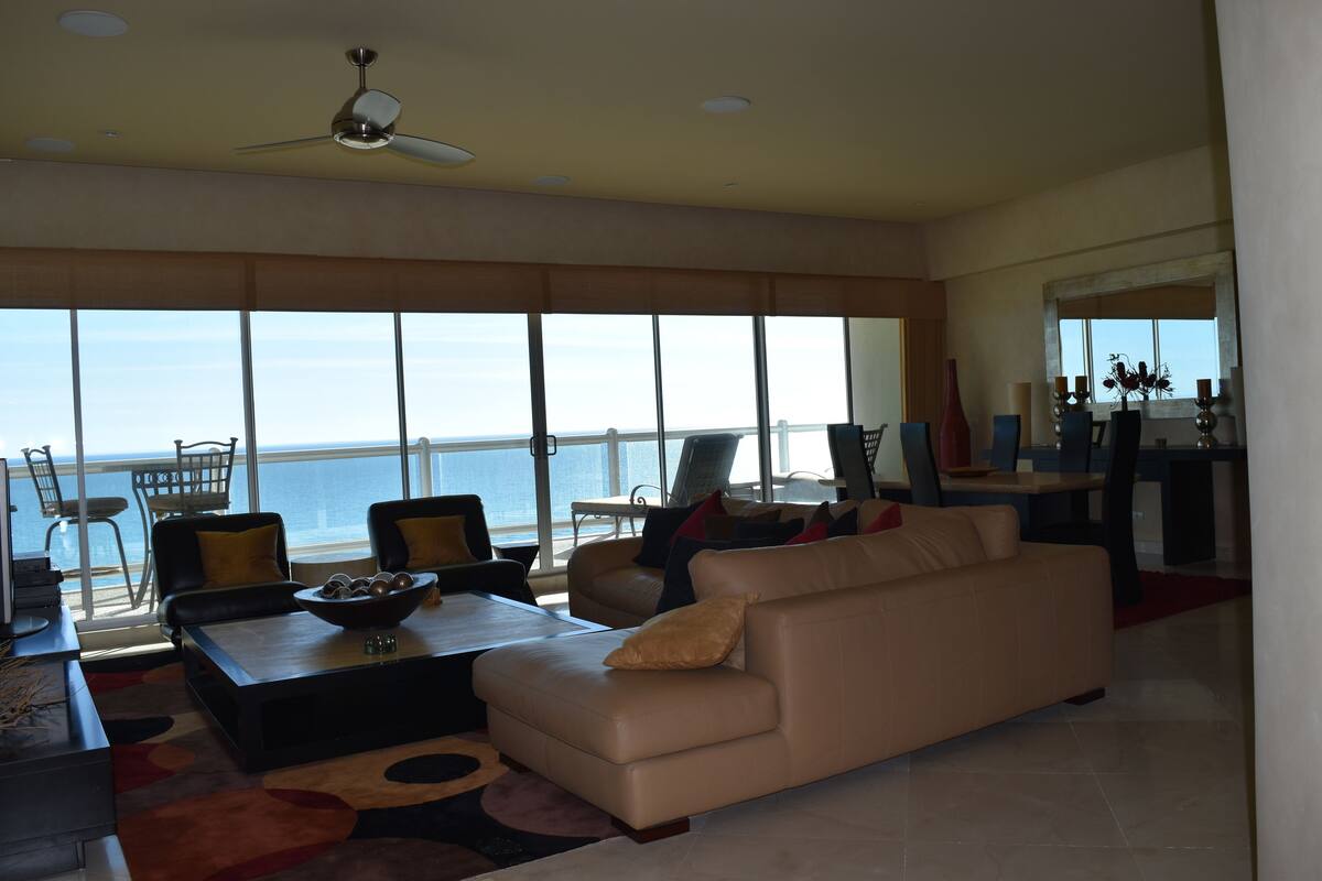 A spacious living room with a large window overlooking the ocean, a ceiling fan, and a dining area with chairs and a table.