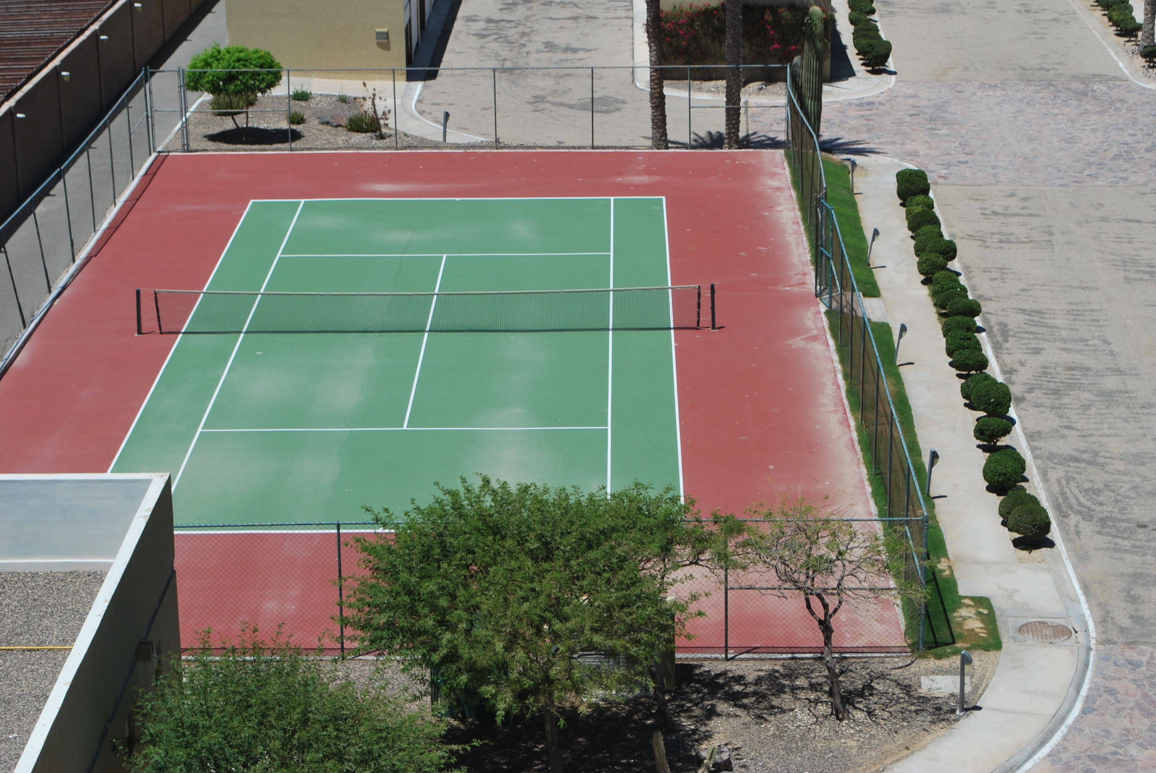 Een tennisbaan met een groene speeloppervlak en een rode omliggende zone, omheind door een metalen hek.