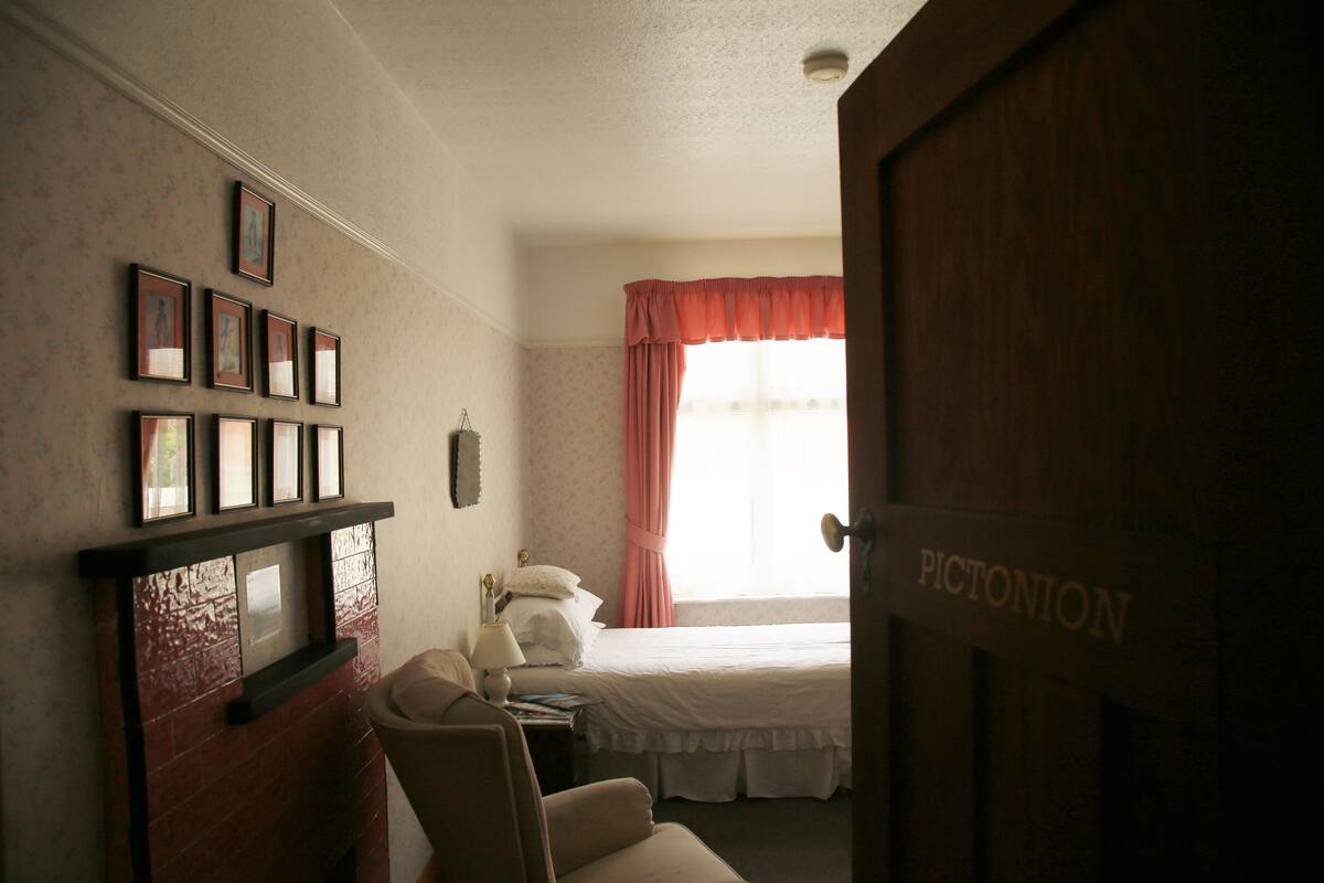 A hotel room with a bed, a chair, a nightstand, a lamp, a shelf with framed pictures, and a door labeled "PICTONION".
