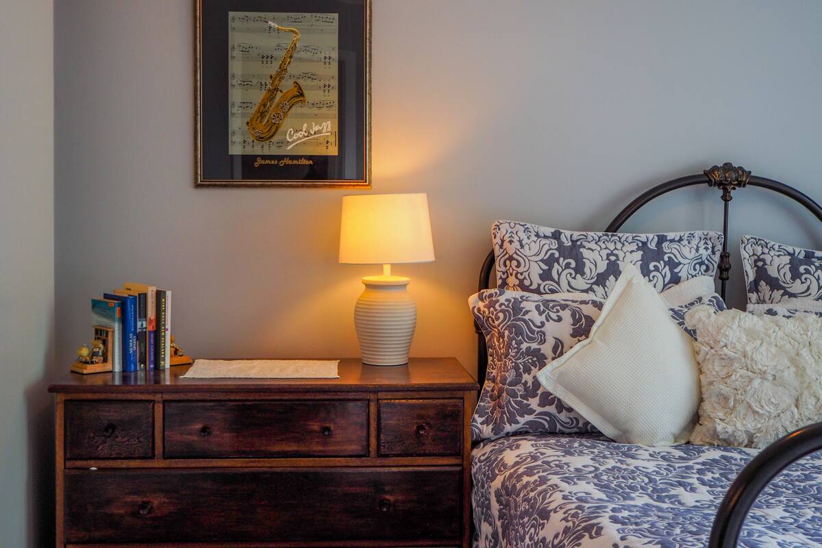 A bedroom with a bed, a bedside table with books, a framed picture, and a lamp.