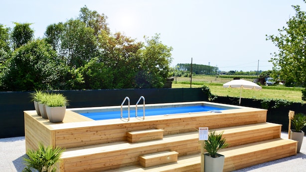 A wooden swimming pool with steps and two ladders, surrounded by potted plants and a clear view of the sky and greenery in the background.