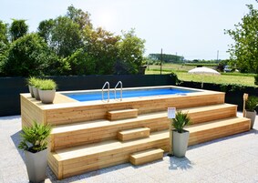 Una piscina in legno con gradini e due scalette, circondata da piante in vaso e con una vista aperta sul cielo e sul verde sullo sfondo.