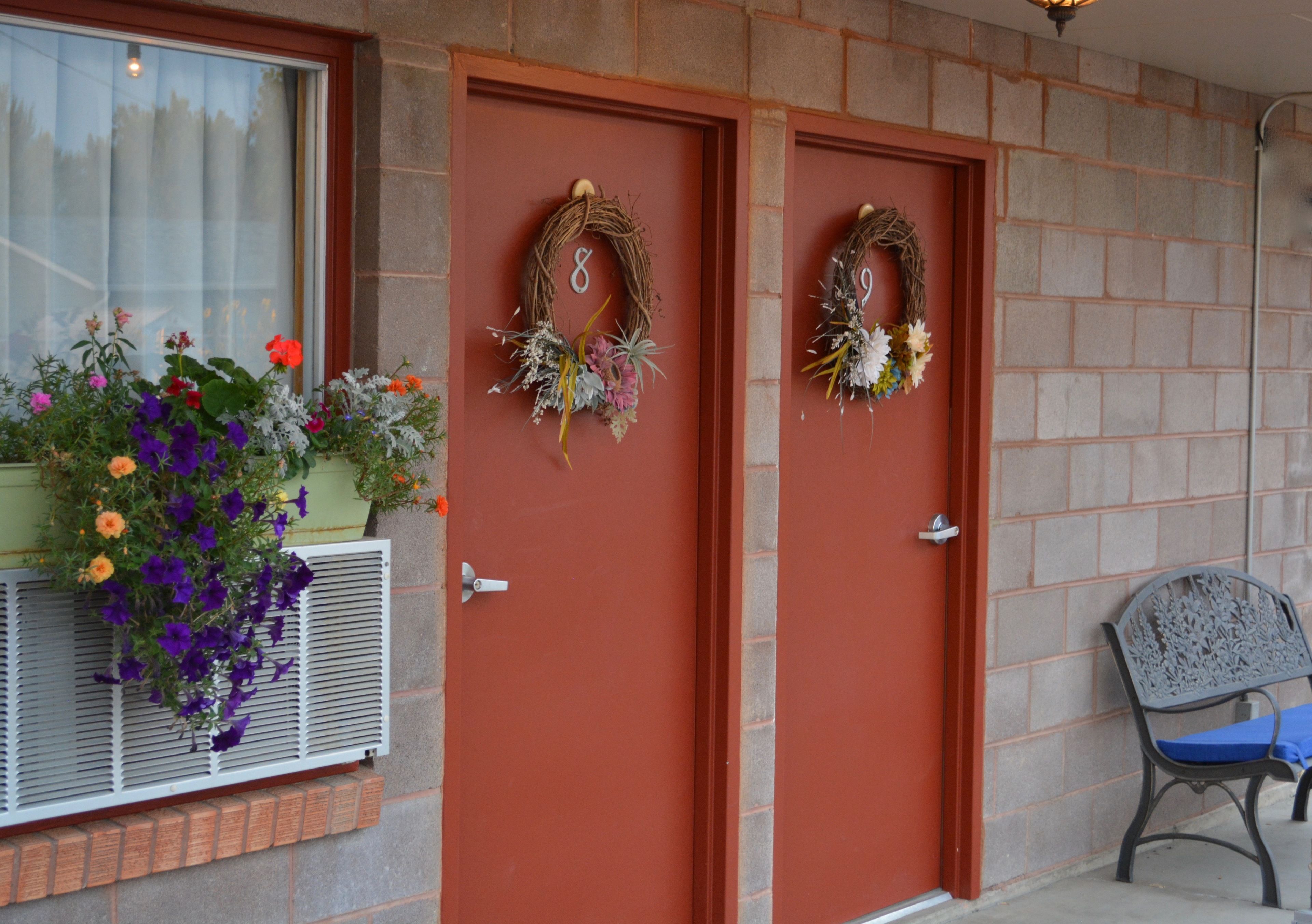 Dos puertas con coronas decorativas, una ventana con un balcón de flores y un banco en el exterior.