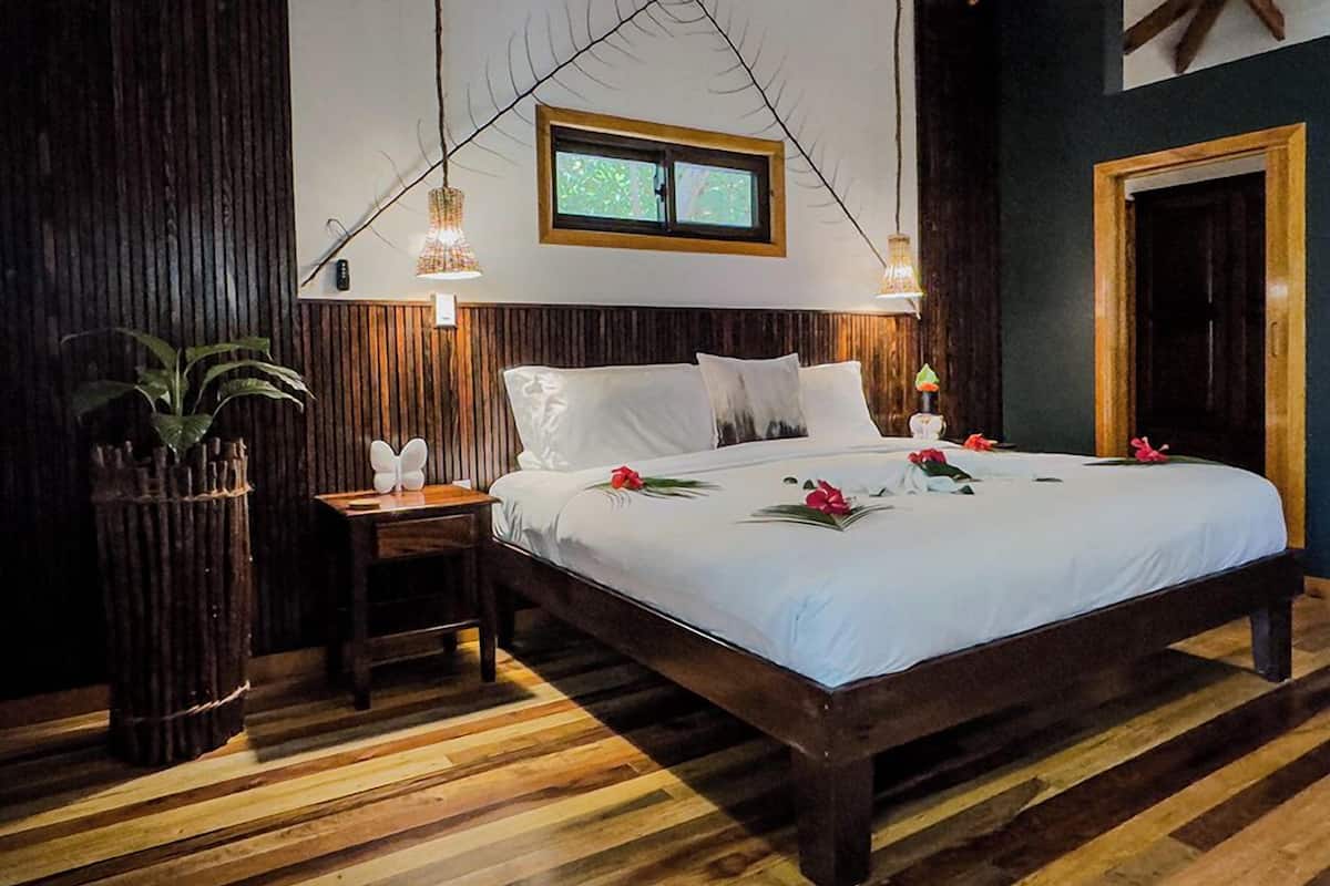 Una cama con estructura de madera, ropa de cama blanca, una mesita de noche pequeña, una planta en maceta y una ventana con vistas a la vegetación.
