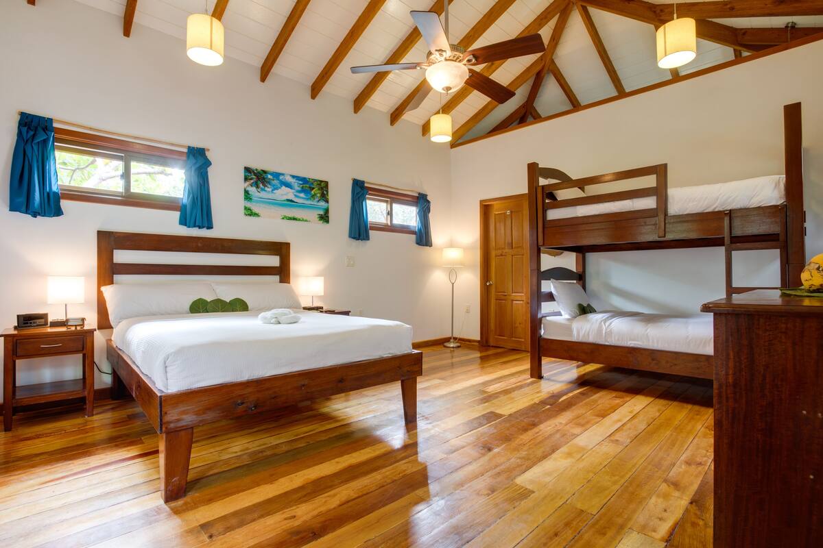 Una habitación con una cama de madera, una cama individual y una litera. Hay ventanas con cortinas, un cuadro en la pared y un ventilador de techo.