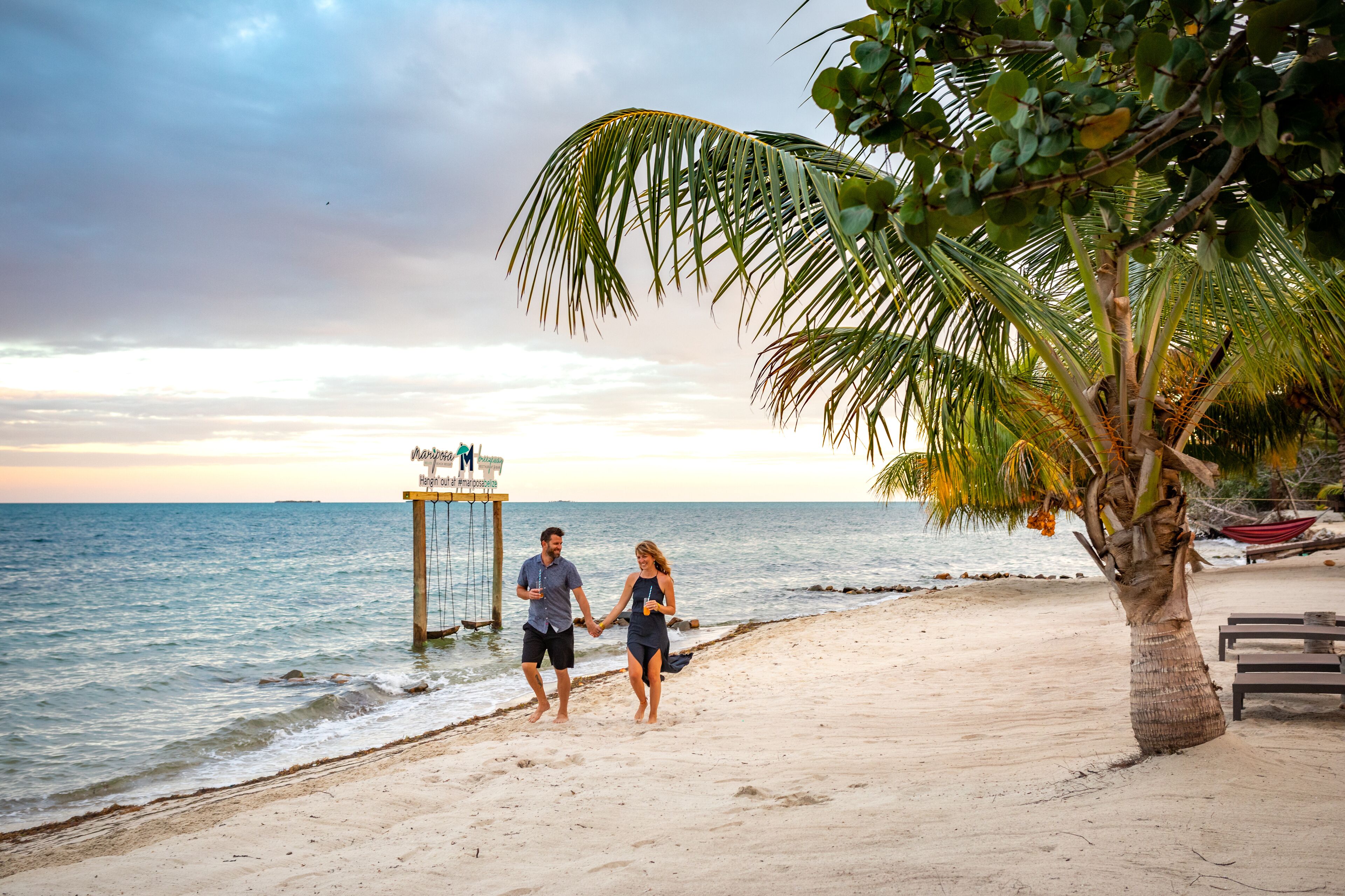 A couple walking on a beach with a palm tree and a sign in the background.
