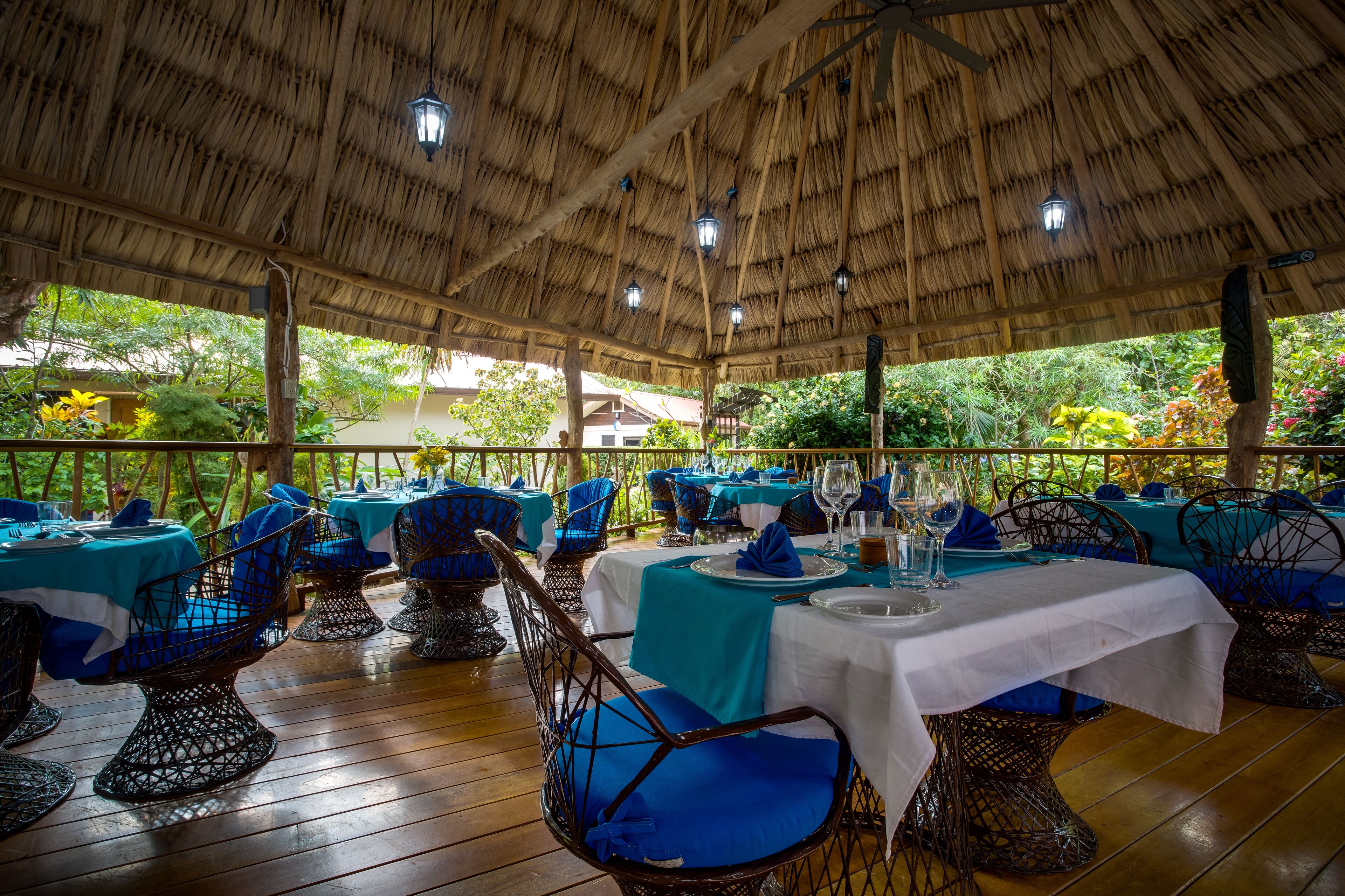 Un restaurante con techo de paja, manteles azules y blancos, sillas de mimbre y vistas a la vegetación.