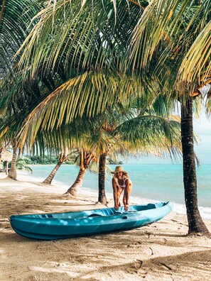 A beach scene with a blue kayak under a palm tree, a person crouching near the kayak, and a clear view of the ocean.