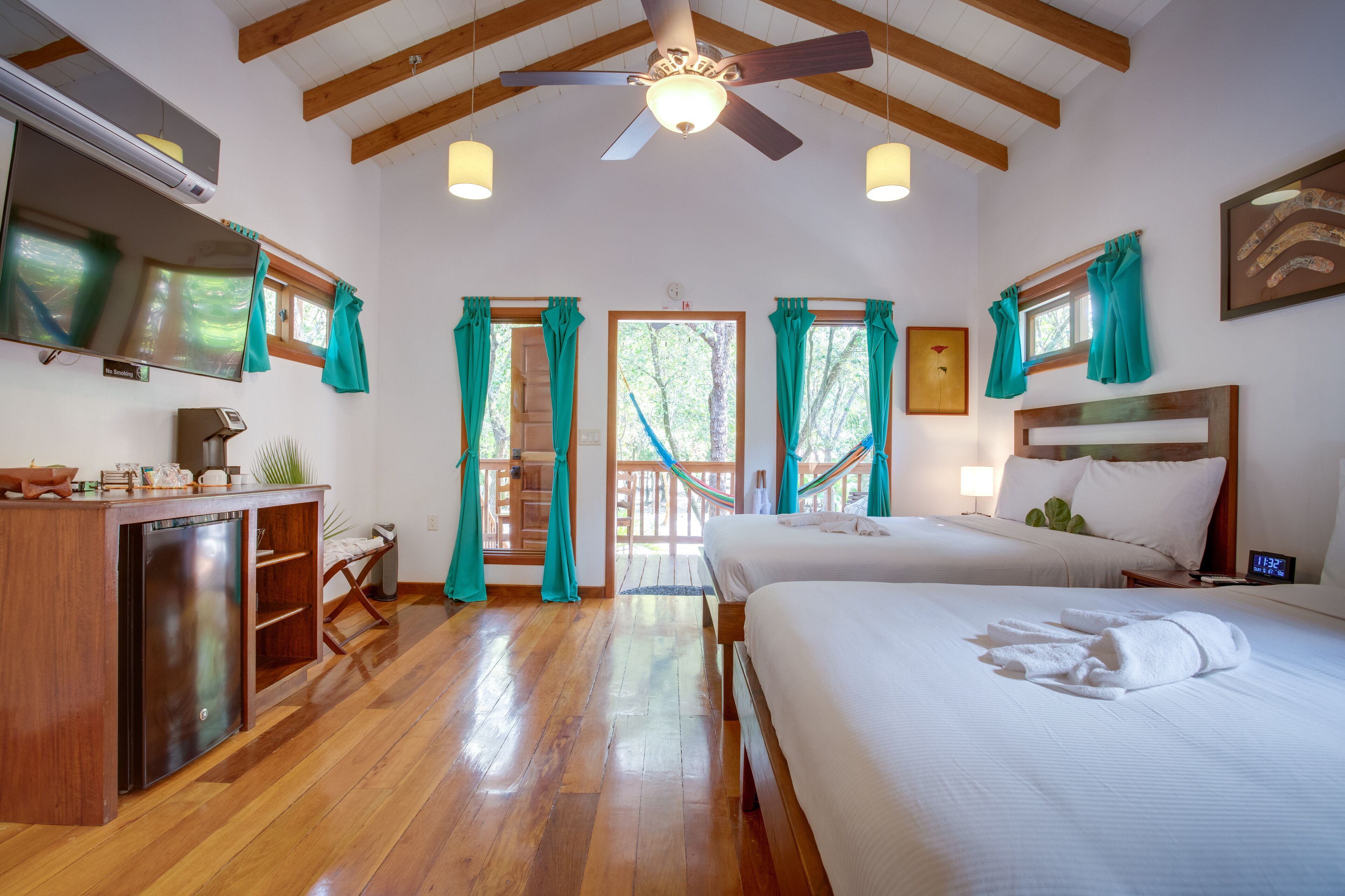 A hotel room with wooden floors, a ceiling fan, a bed, a desk with a computer, and a TV.