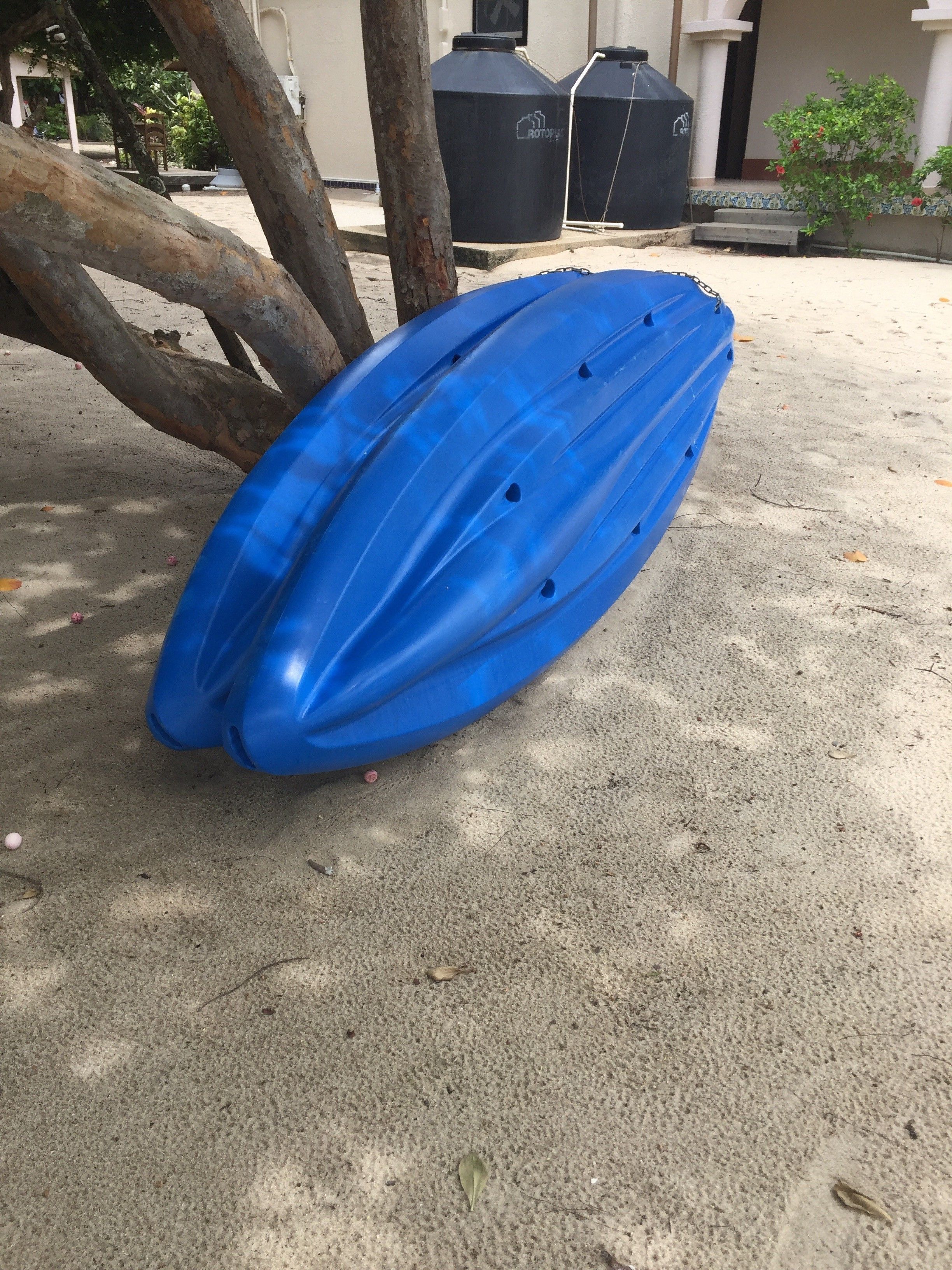 Un kayak azul reposa sobre arena, con un árbol y contenedores negros al fondo.