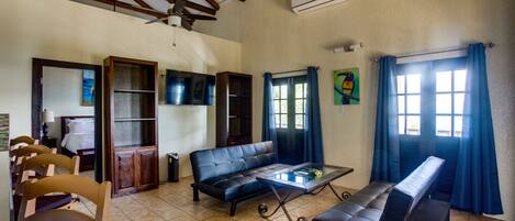 A spacious living area with a sofa, a coffee table, and a bench. There is a television mounted on the wall, and a ceiling fan is visible.