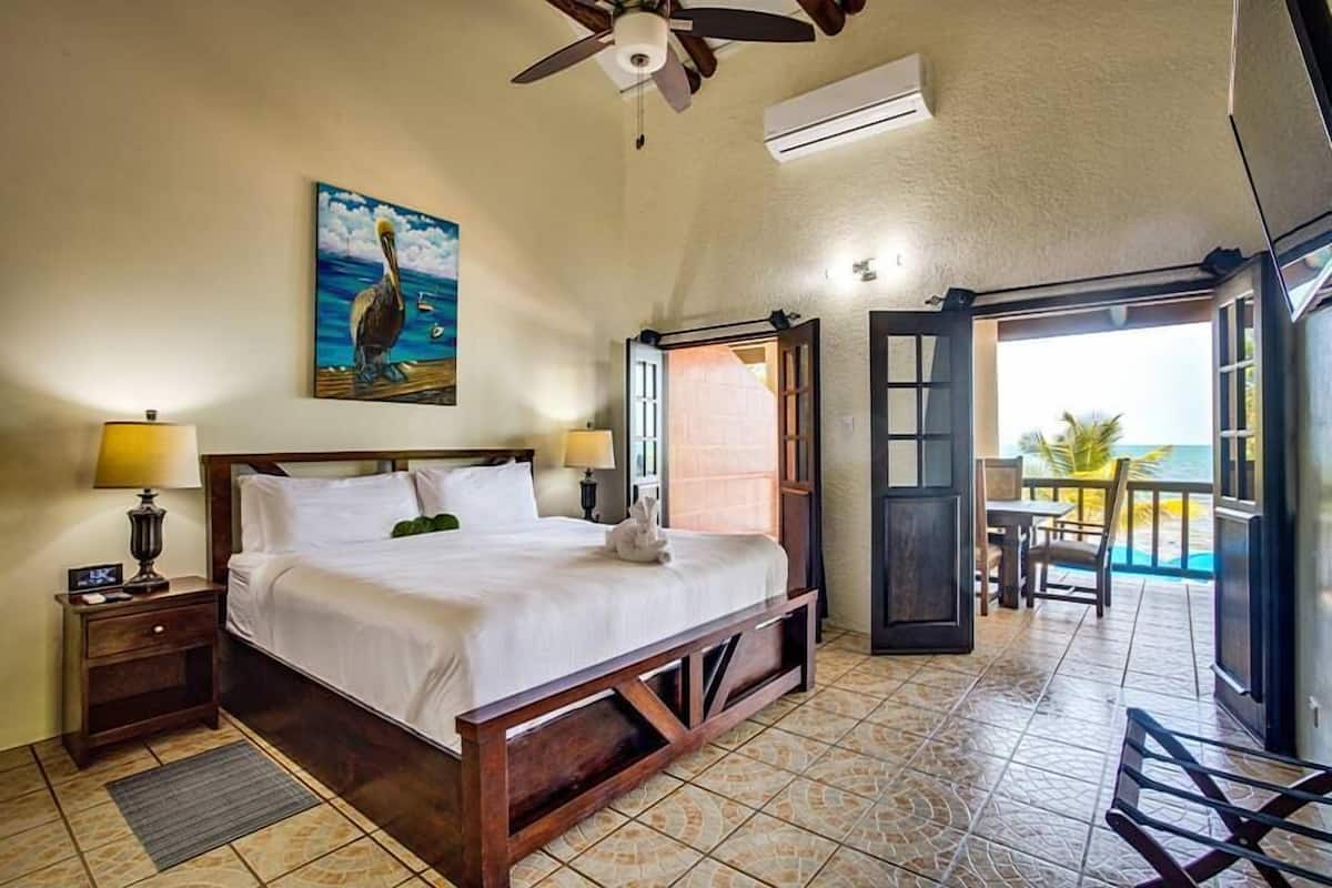 Un dormitorio con cama, mesita de noche, lámpara, un cuadro de un pelícano y una puerta abierta que da a un balcón con vistas al océano.