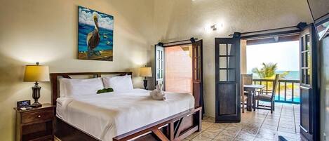 A bedroom with a bed, a nightstand, a lamp, a painting of a pelican, and an open door leading to a balcony with a view of the ocean.