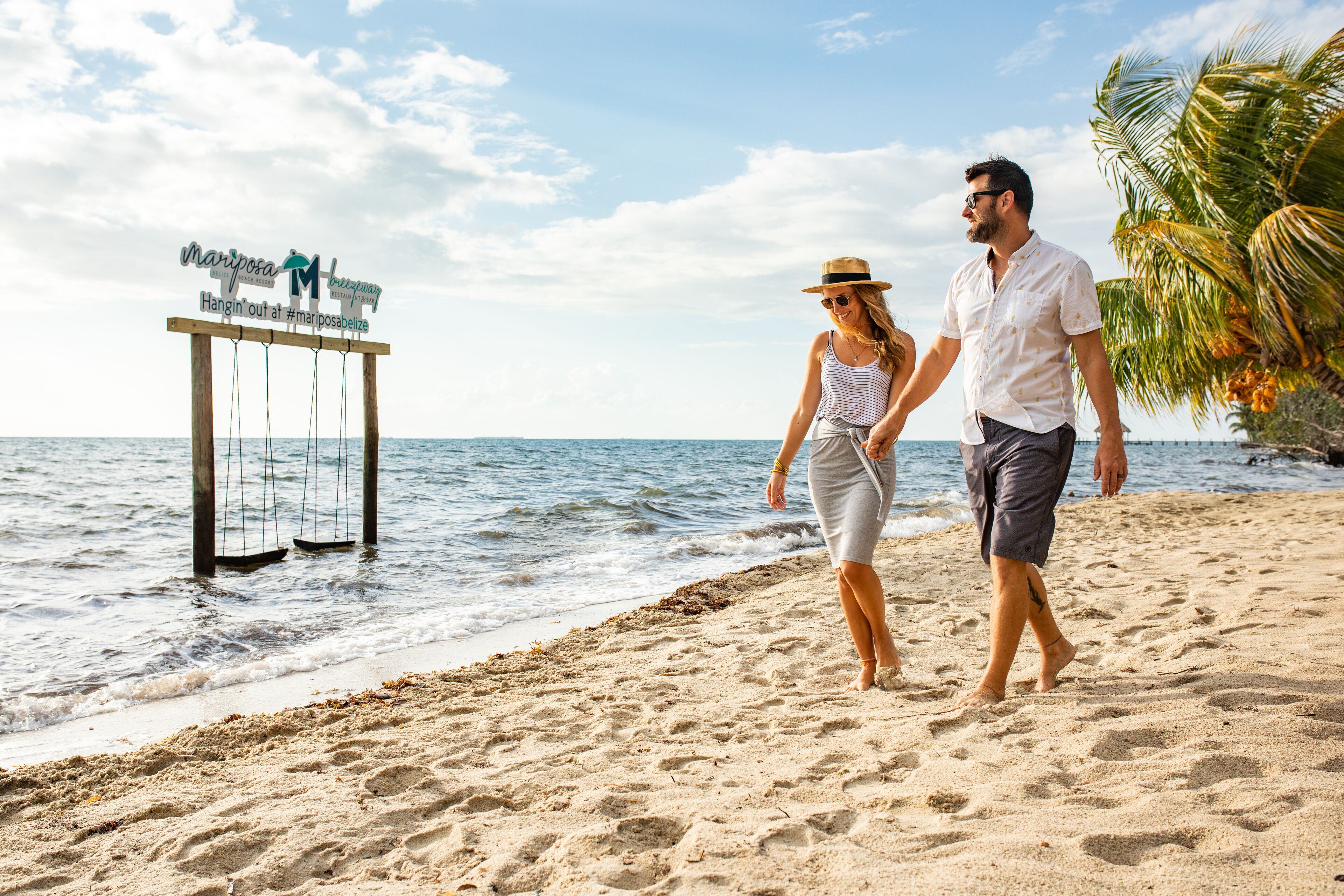 A couple walking on a sandy beach with a signboard in the background.