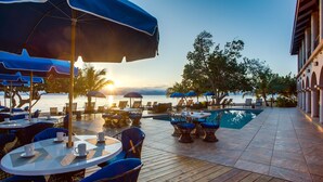 A poolside area with tables and chairs, umbrellas, and a view of the sea at sunset.