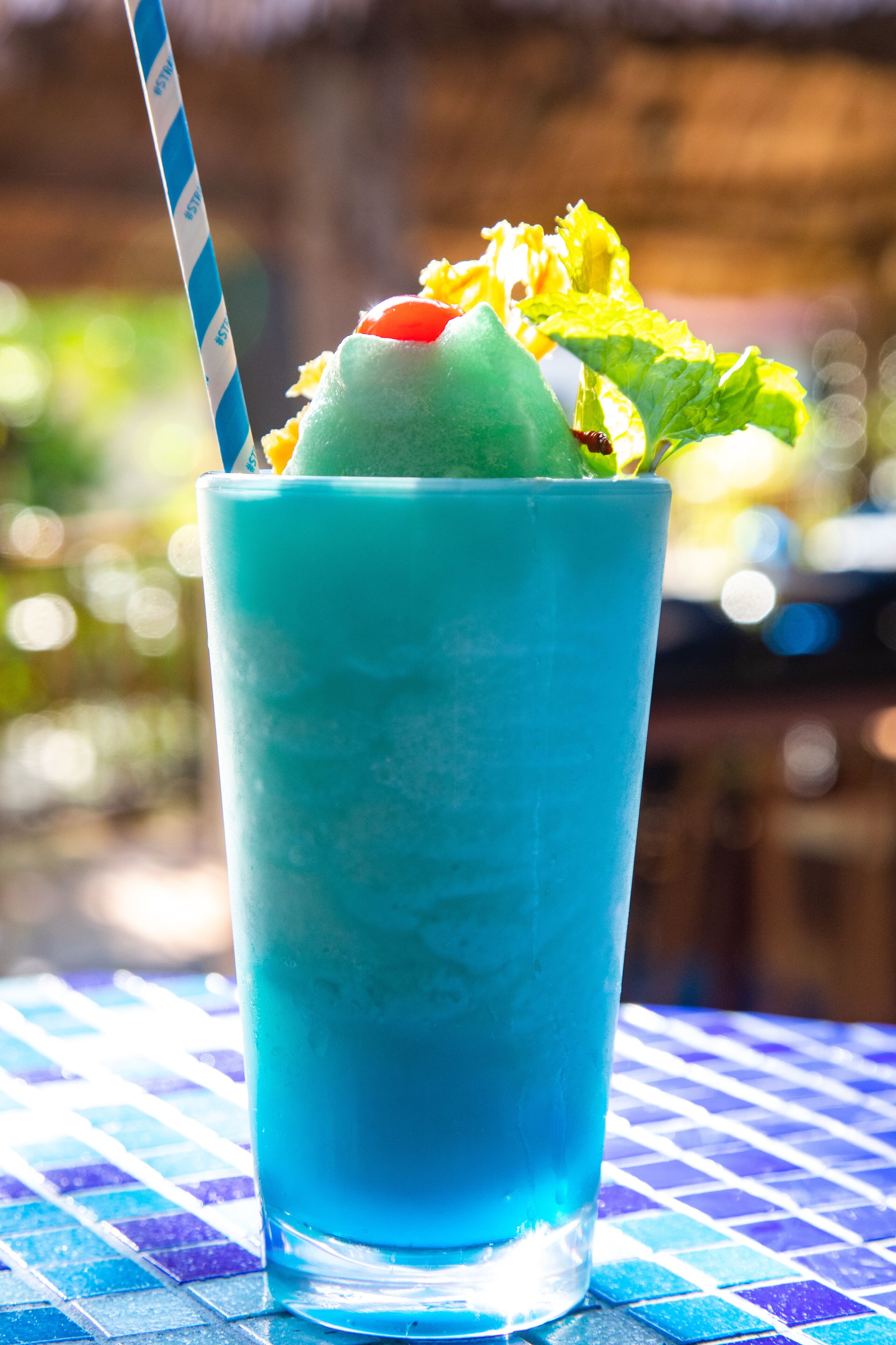 A tall glass with a blue beverage, a straw, and a green scoop on top, placed on a patterned table.