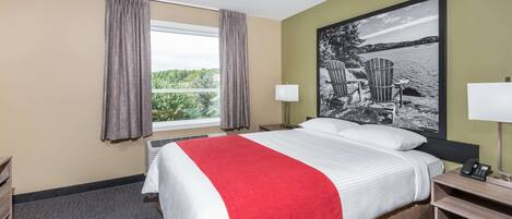A hotel room with a large bed, a window with curtains, a desk with a lamp, and a painting on the wall.