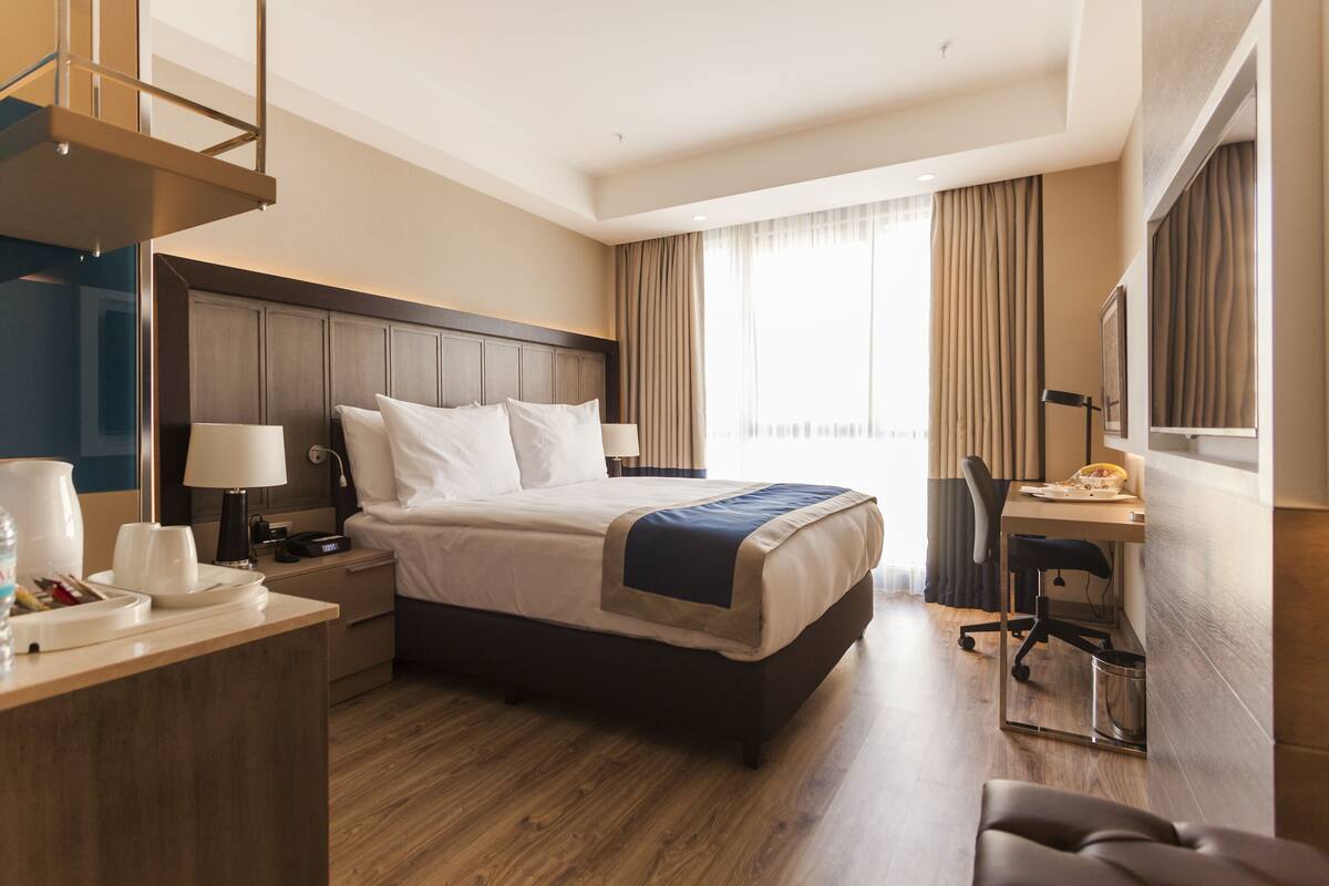 A hotel room with a bed, desk, chair, and a small table with a lamp.