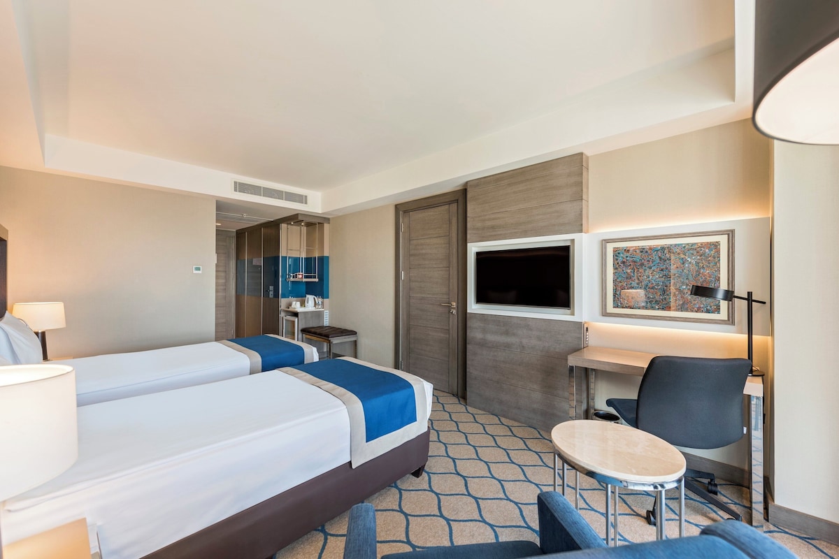 Een hotelkamer met twee bedden, een bureau, een stoel, een kleine tafel, een lamp, een televisie en een schilderij aan de muur.