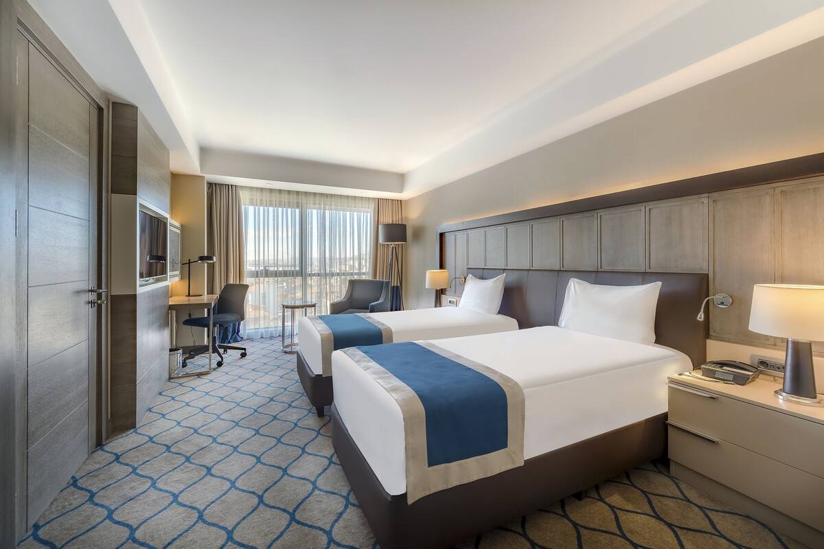 A hotel room with two beds, a desk, a chair, a lamp, and a large window with curtains.