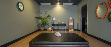 A room with a large black exercise mat, a potted plant, a wall clock, and a mirror with hexagonal frames.