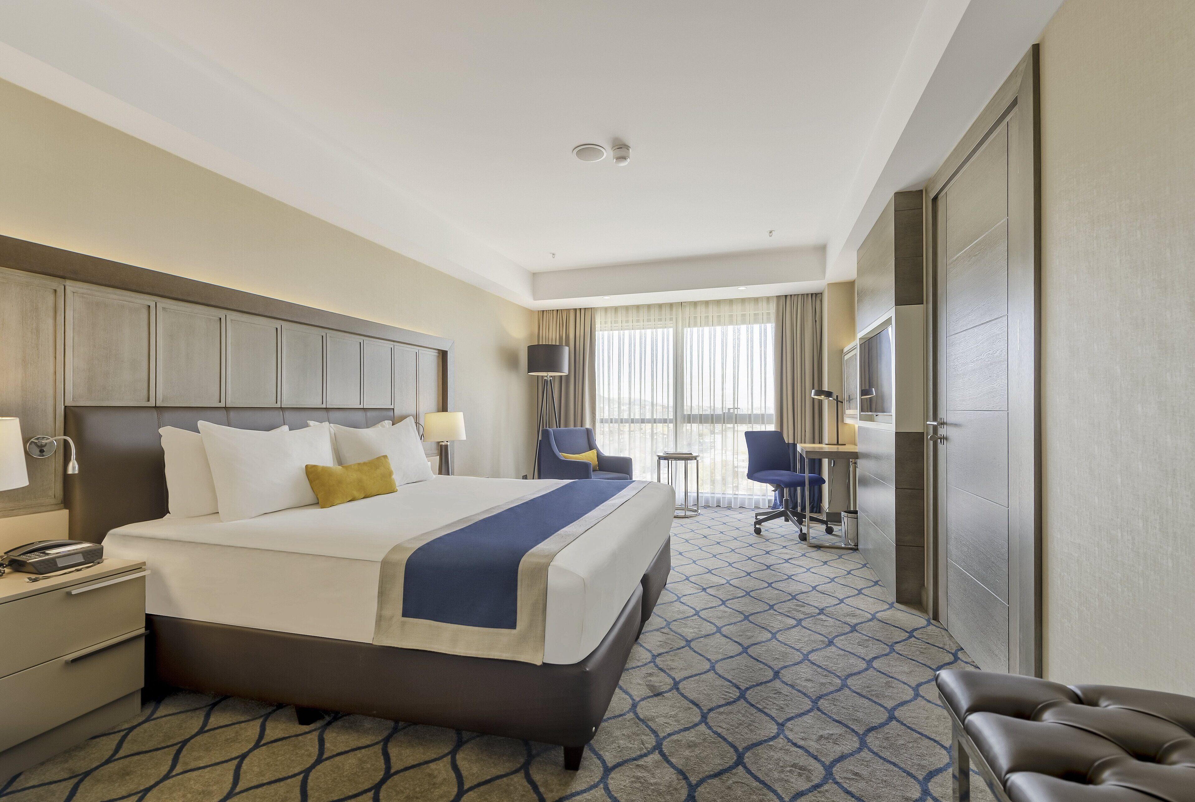 A hotel room with a large bed, bedside tables, a desk, a chair, a TV, and a wardrobe.