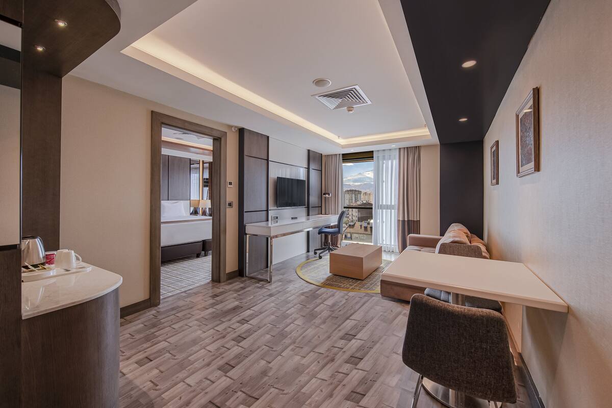 A modern hotel room with a kitchenette, a seating area, a large window with a city view, and a flat-screen TV.