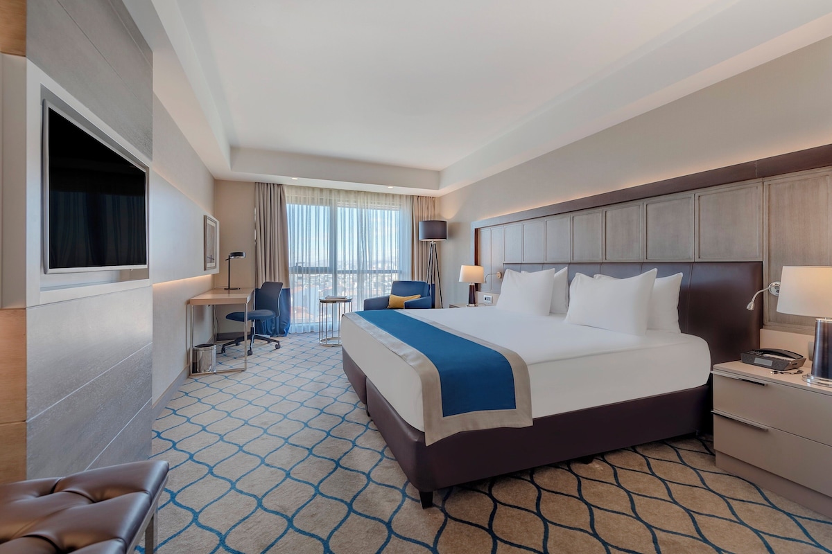 Een hotelkamer met een groot bed, een bureau met stoel, een televisie en een raam met gordijnen.